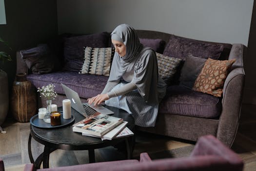 A Muslim woman in traditional attire working on her laptop in a cozy living room setting.
