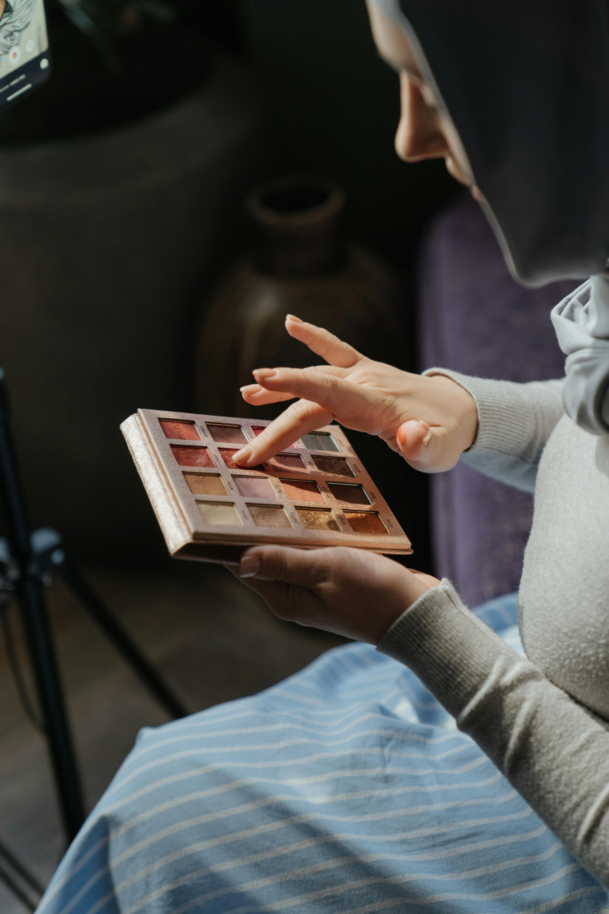 A woman in a hijab applies eyeshadow using a palette indoors, highlighting a beauty routine.