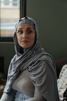 Thoughtful woman wearing hijab and glasses, sitting in a cozy indoor setting.