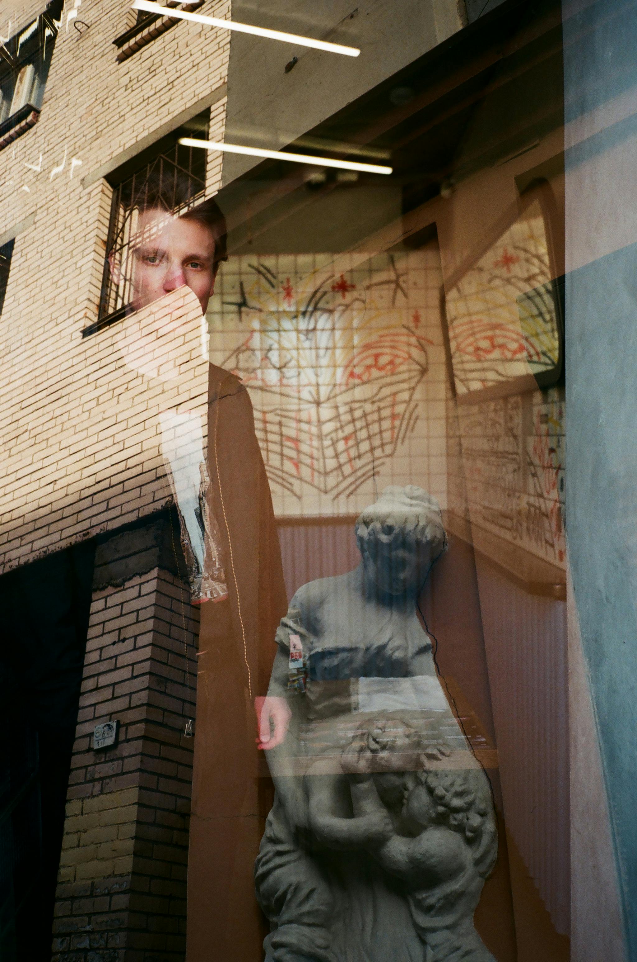 Through glass of serious young male looking at camera while standing in dark room with statue behind transparent window with reflection of multi storey brick house on surface of glass