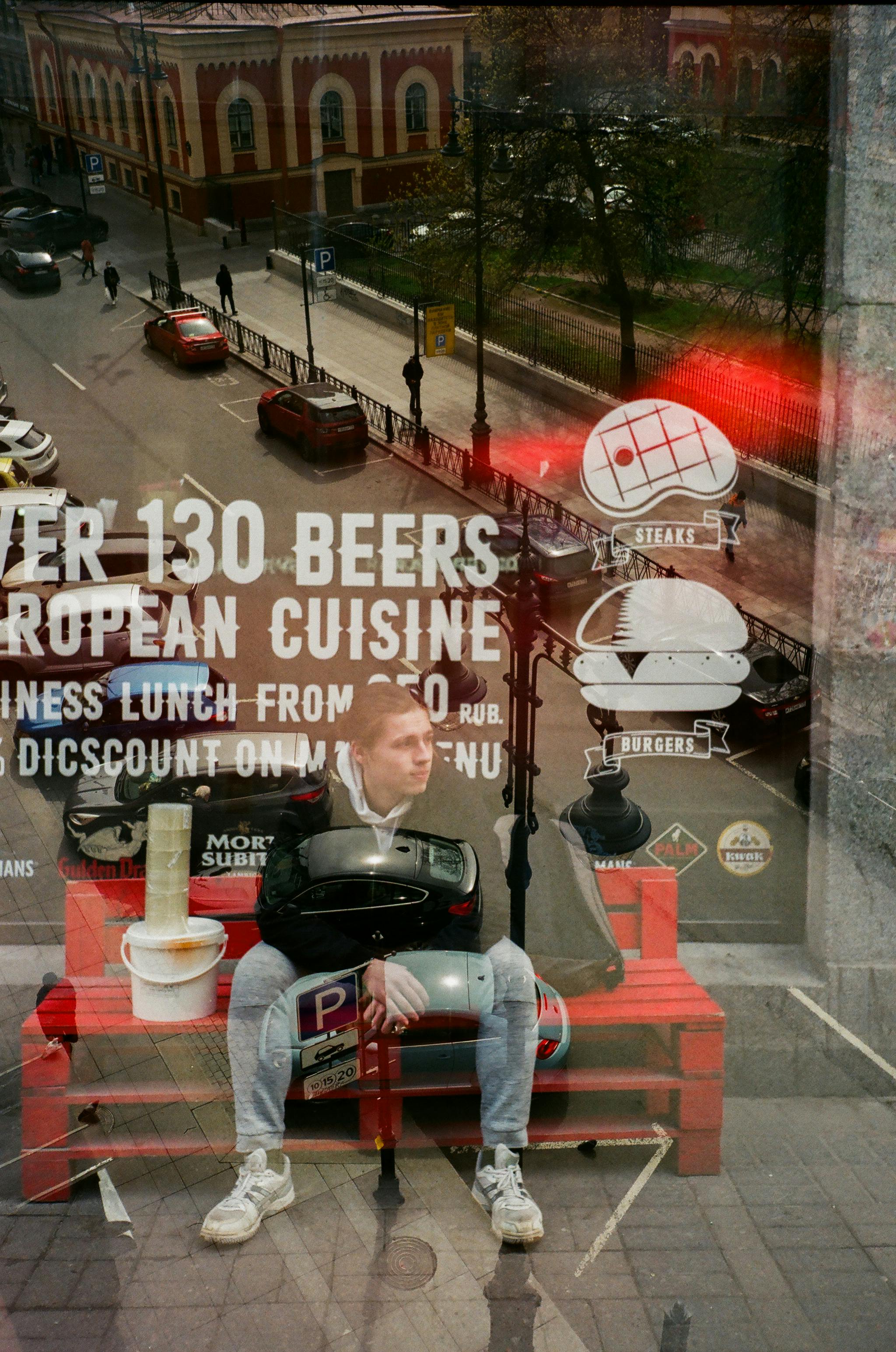 Through glass of young pensive man in casual clothing looking away while resting on red wooden bench