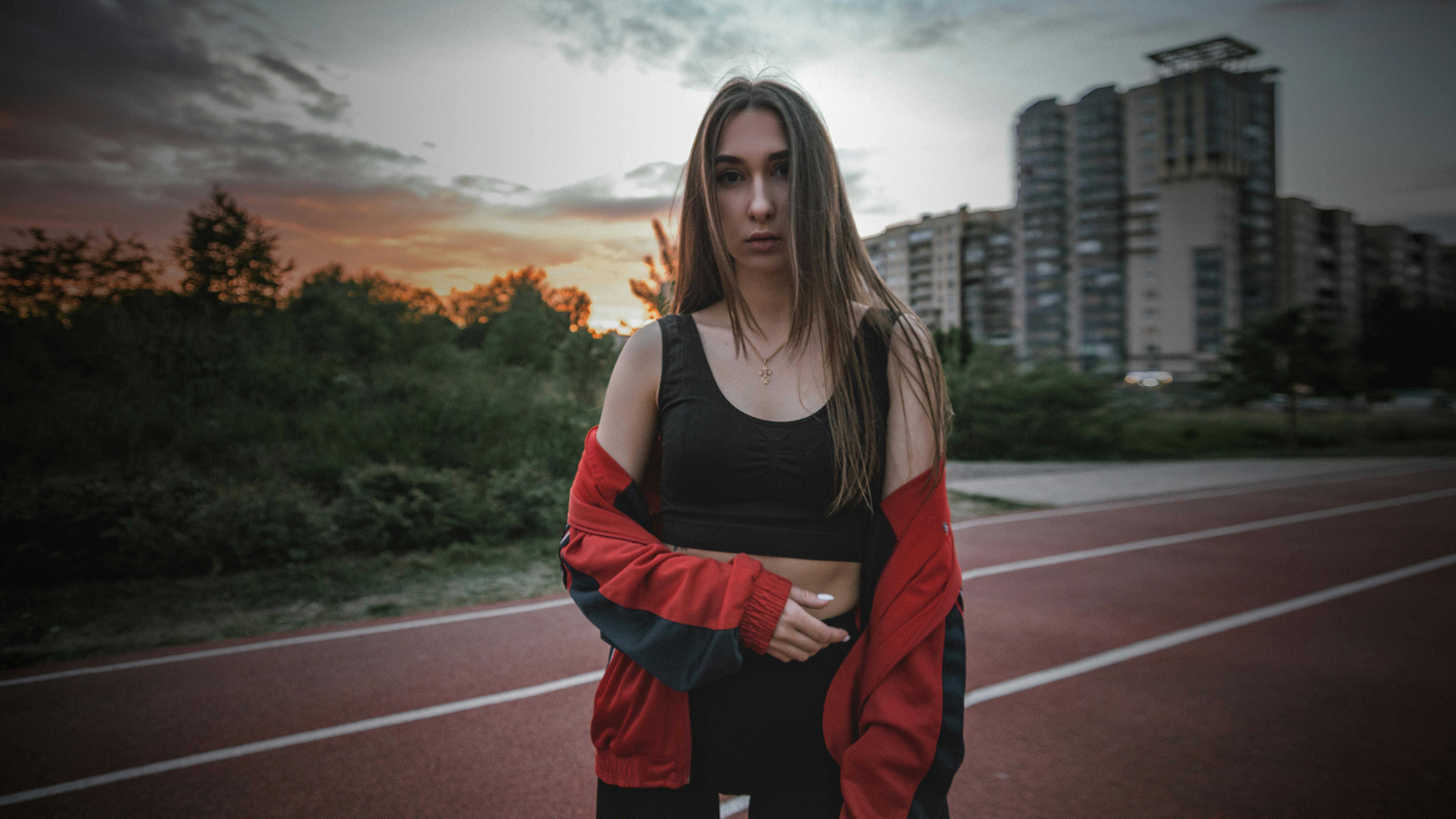 A fashionable woman in a sports bra and jacket poses on a track at sunset.