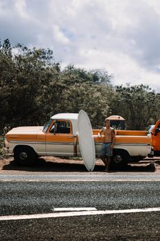 Shirtless surfer with surfboard next to retro pickup truck, roadside scene.