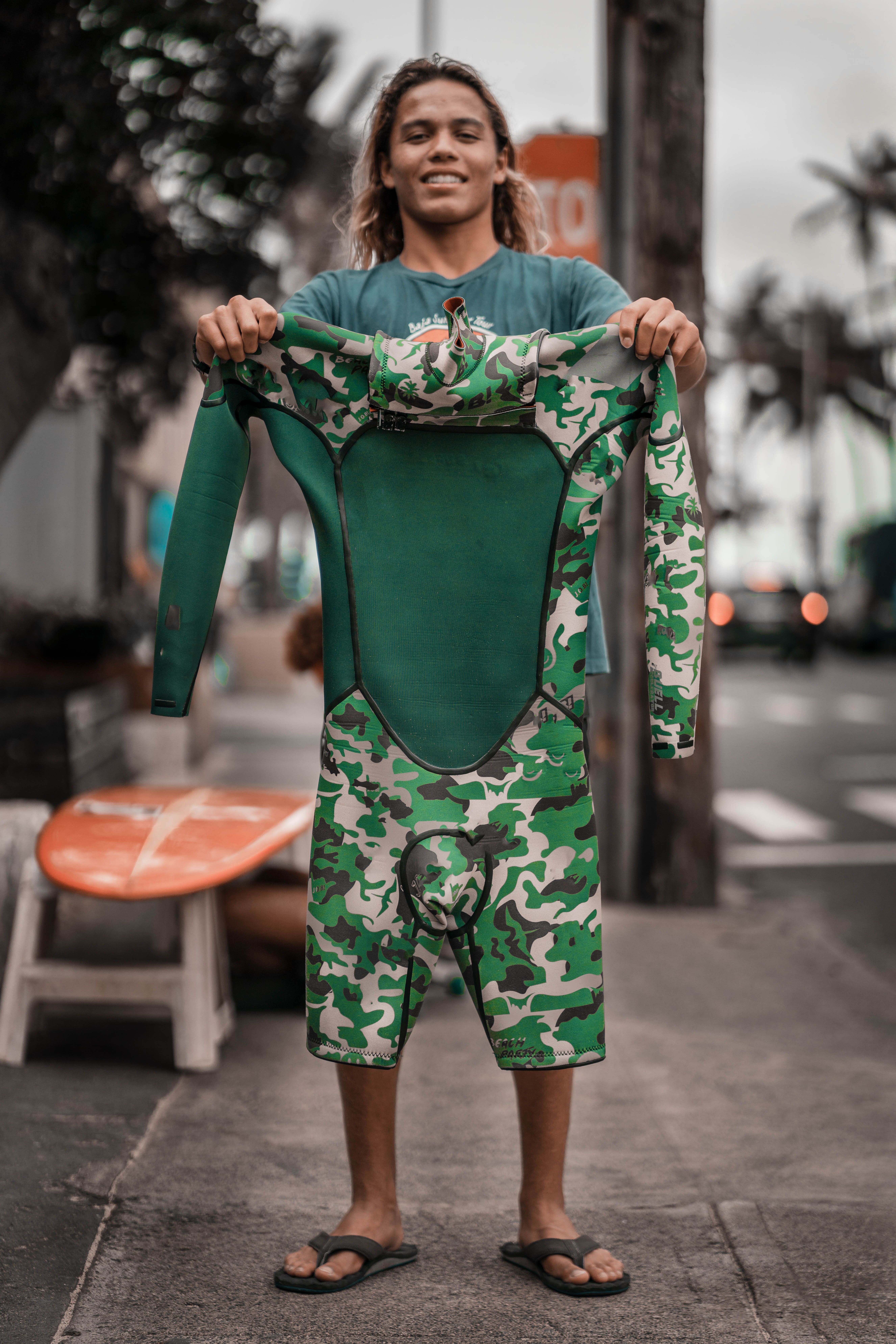 A man holds a camo wetsuit on a city street, with a surfboard in the background.