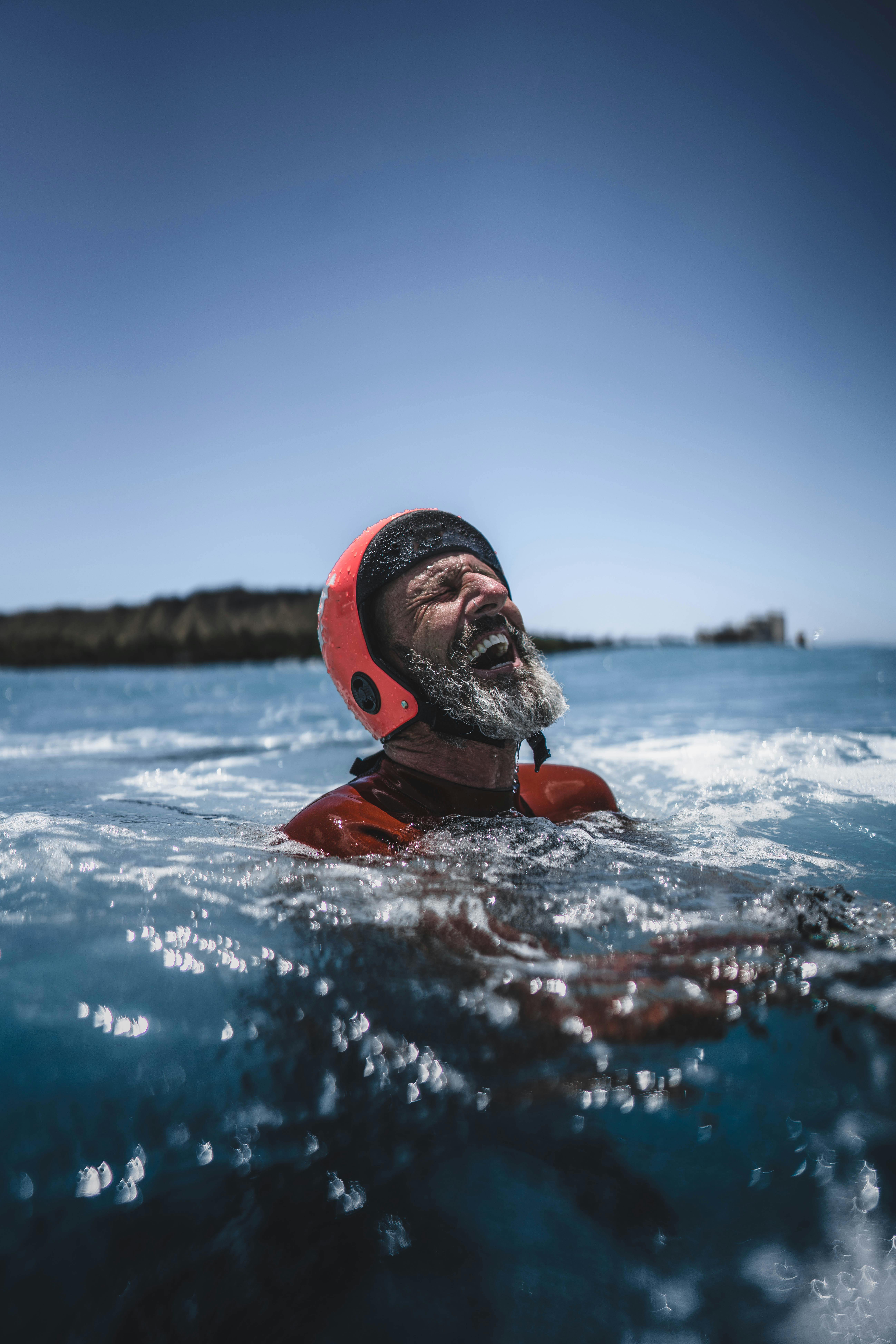Elderly man enjoying a swim in the ocean wearing a protective helmet under the bright sun.