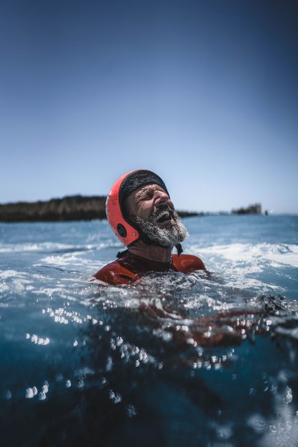 Elderly man enjoying a swim in the ocean wearing a protective helmet under the bright sun.