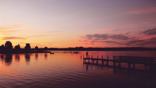 Free A tranquil scene of a lone person standing on a dock at sunset with vibrant skies reflecting on the water. Stock Photo
