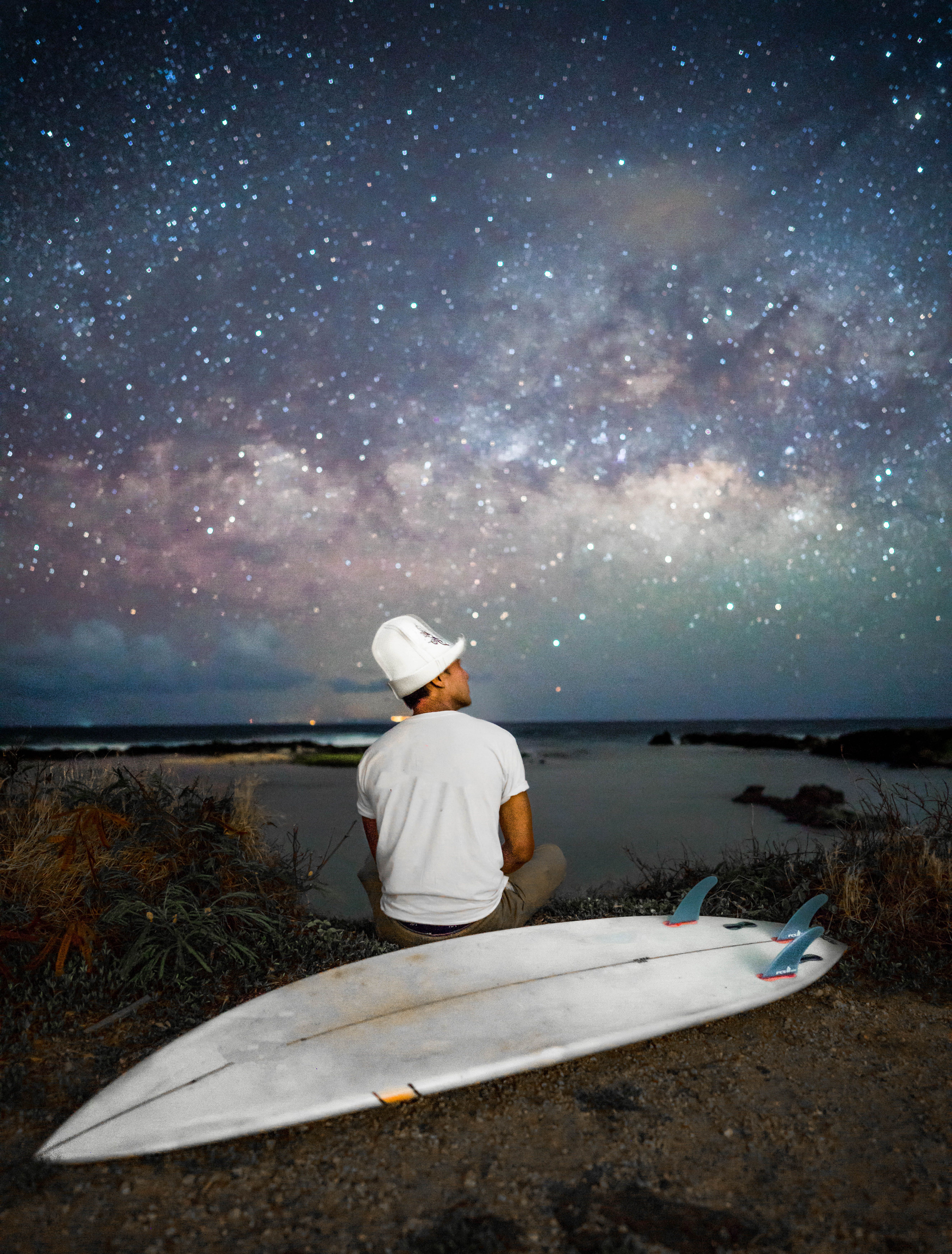 一名孤独的男子坐在海滩上，凝视着星空，身旁放着他的冲浪板