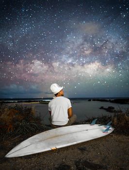 一名孤独的男子坐在海滩上，凝视着星空，身旁放着他的冲浪板