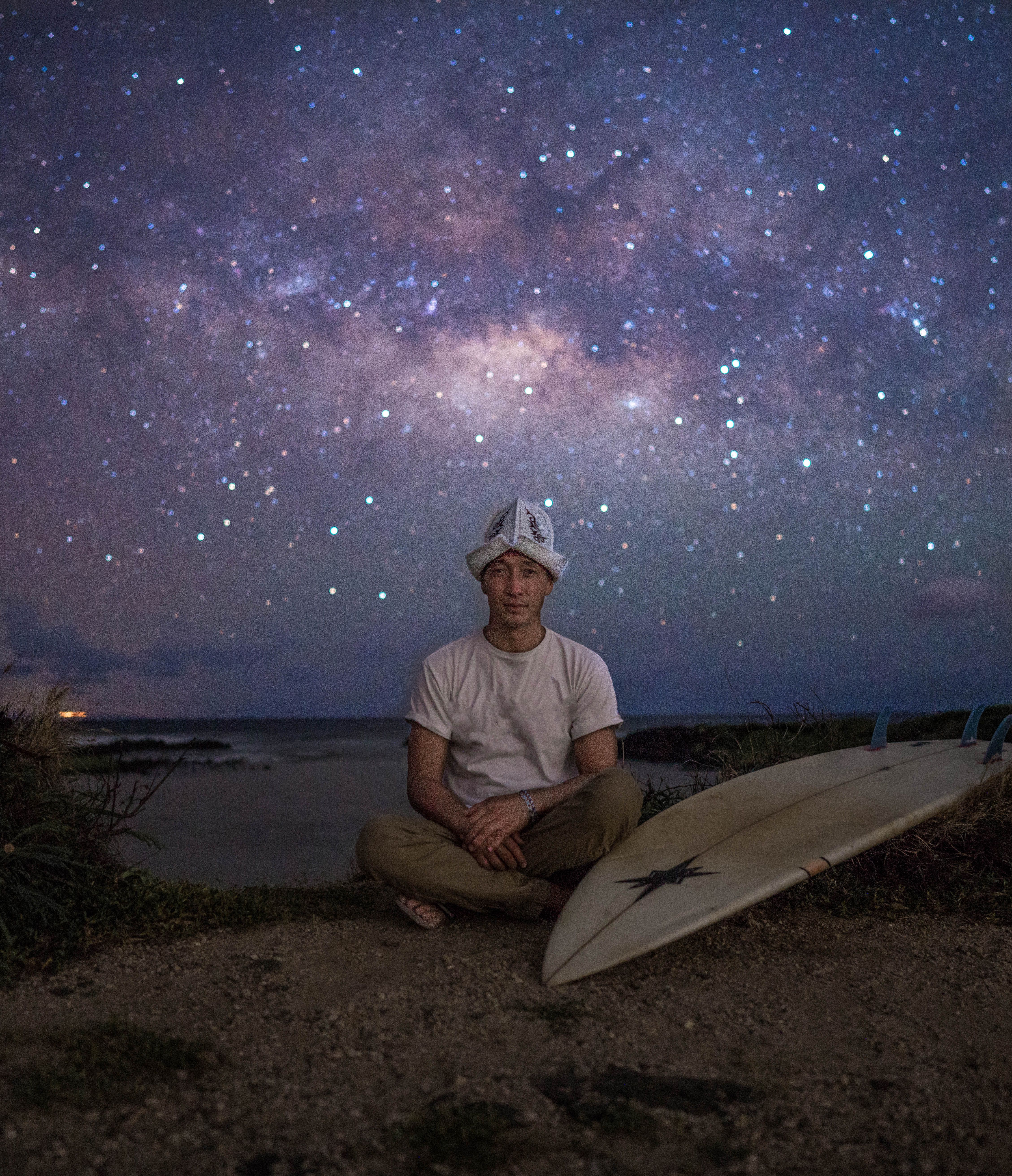 A man with a hat sits beside a surfboard, gazing at a star-filled night sky by the coast.