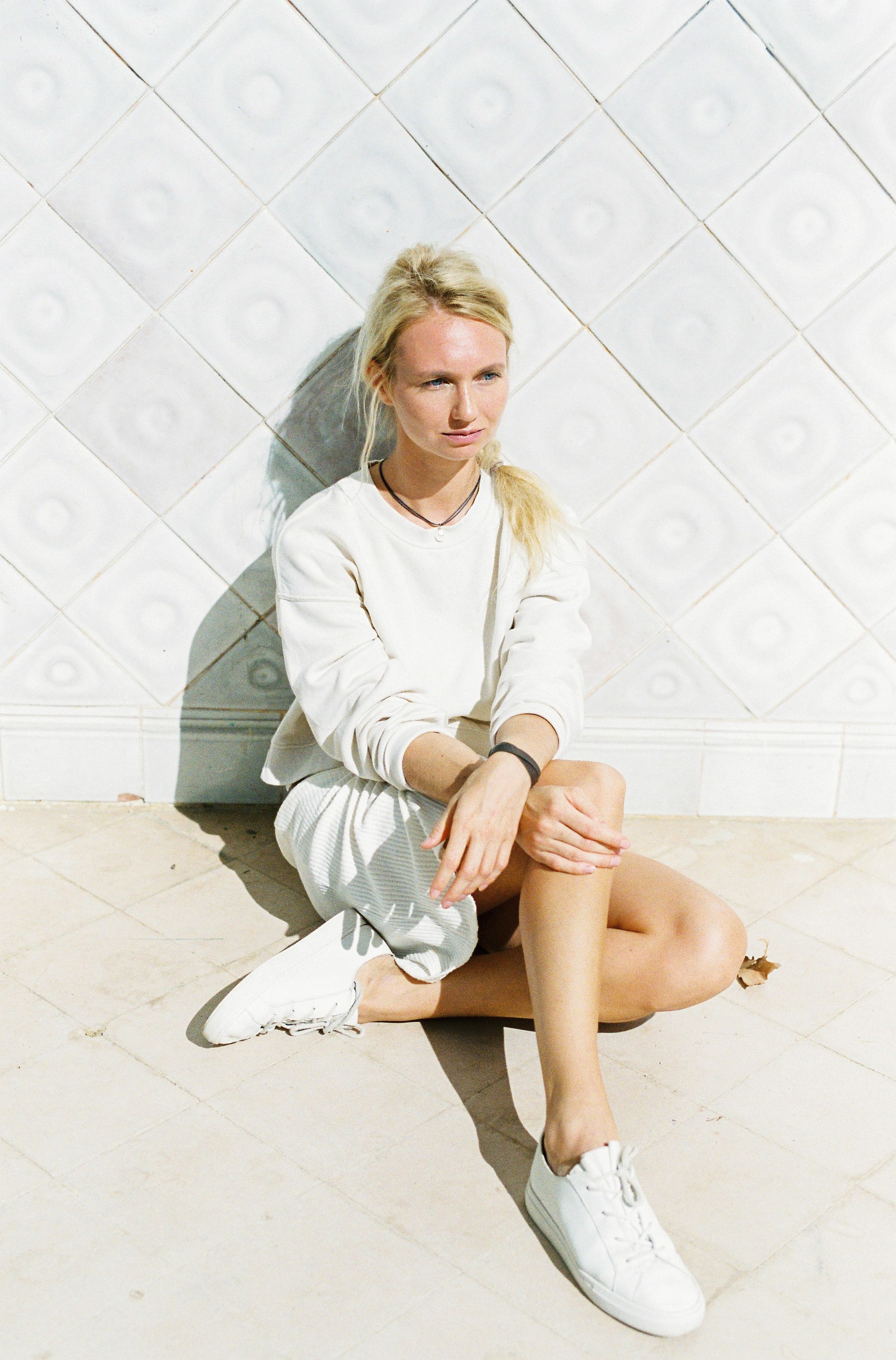 A blonde woman sitting casually against a tiled wall on a sunny day.
