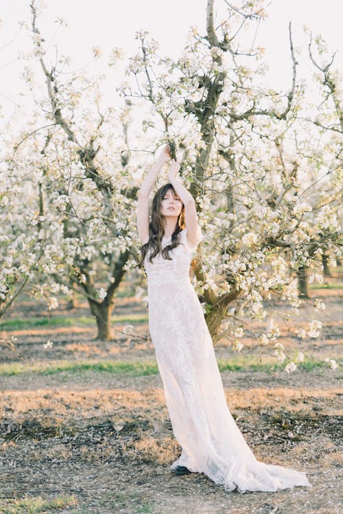 Bride in Wedding Dress Standing with Arms Raised in Orchard