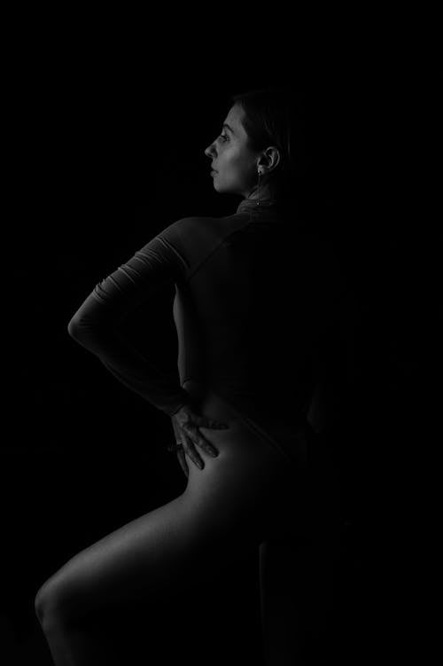 Black and white of young pensive female in body suit with long sleeves looking away on black background
