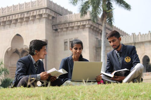 Free Ground level of young ethnic students in uniform sharing laptop and books while preparing for exam in yard of university Stock Photo