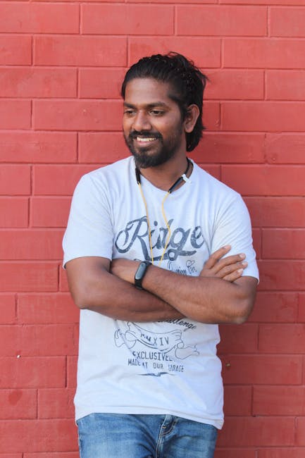 A cheerful man with a beard stands casually against a red brick wall, exuding confidence.