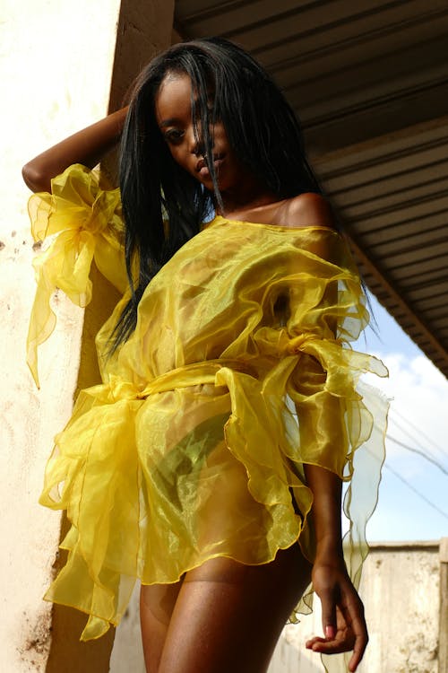 Free From below of serious young African American female in short transparent yellow dress looking at camera while touching long dark hair Stock Photo