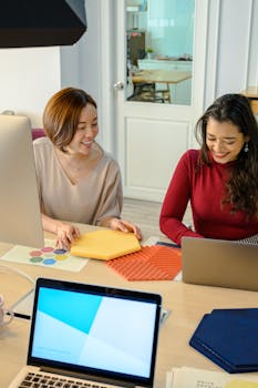 Two women working and smiling at desks with laptops in a modern office.