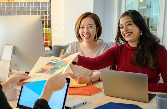 Two women colleagues exchanging ideas in a bright, modern office, working on laptops and documents.