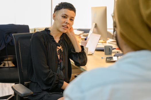 A woman in a black dress engages in a conversation while sitting at a desk in a modern office.