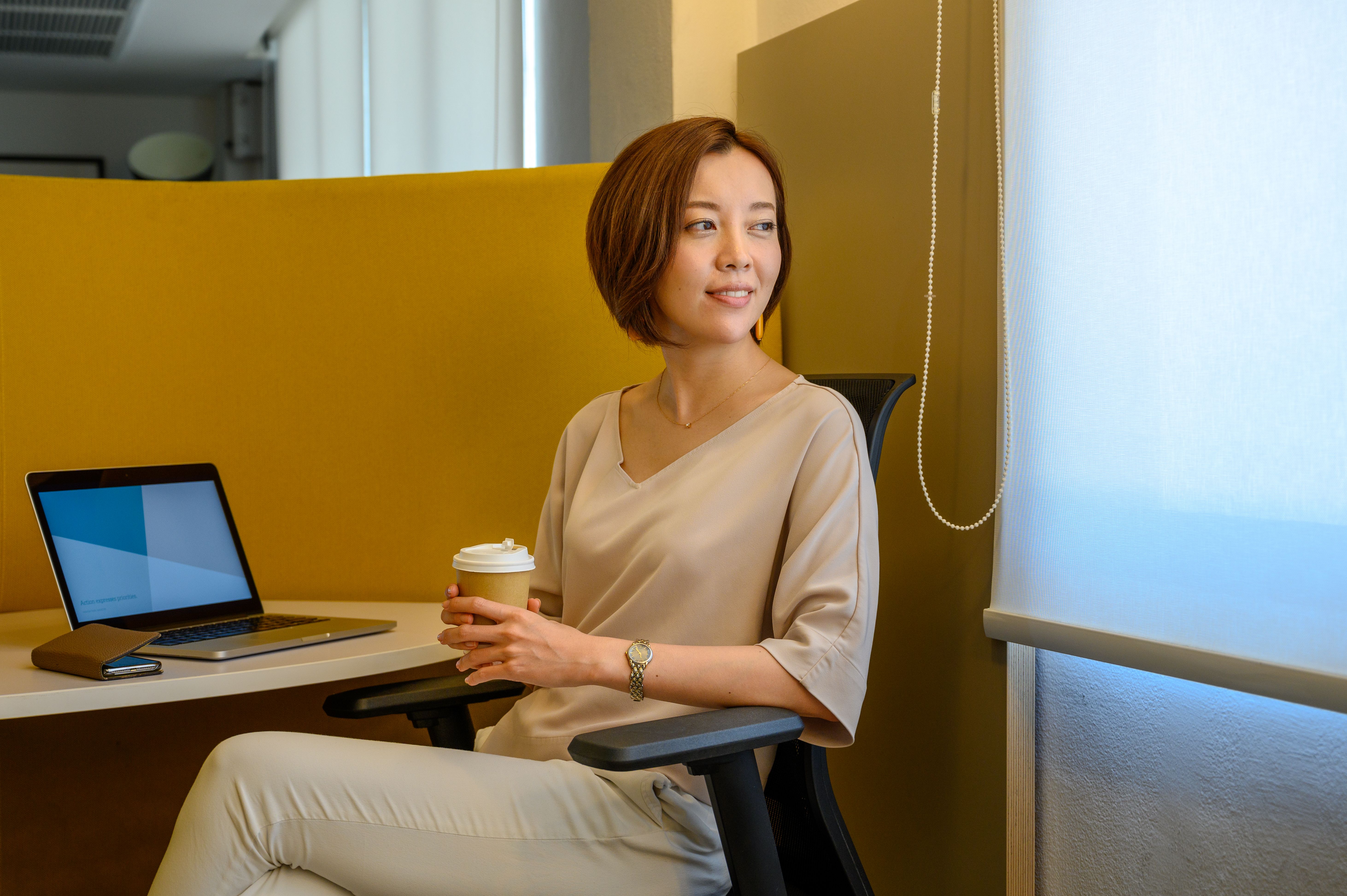 一位女士坐在办公椅上，手拿咖啡杯，旁边放着笔记本电脑