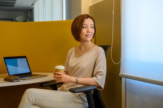 一位女士坐在办公椅上，手拿咖啡杯，旁边放着笔记本电脑