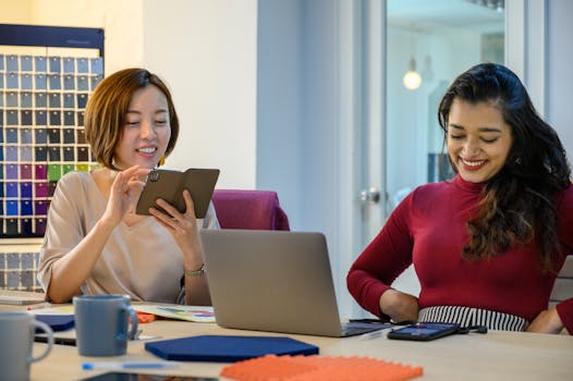 Two women collaborating in a modern office environment, using laptops and smartphones.