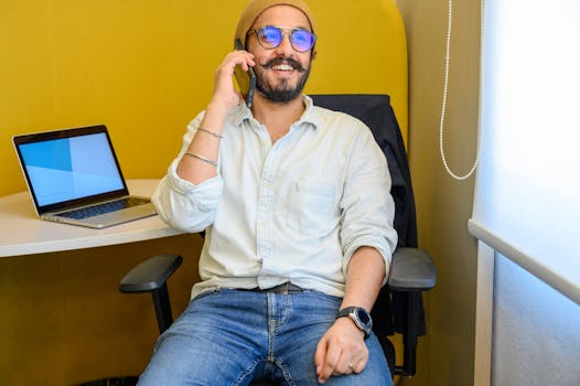 Bearded man with glasses talking on smartphone, seated on office chair beside laptop.