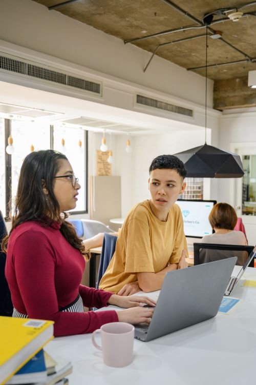 Free Two colleagues engaging in a conversation at a shared workspace, fostering teamwork and collaboration. Stock Photo
