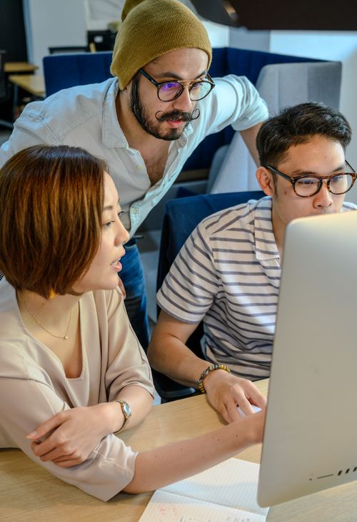 Free Three young adults collaborating on a project at a desk with a computer monitor. Stock Photo
