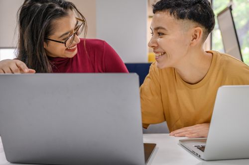 Free Two diverse colleagues smiling and collaborating on laptops in an office setting. Stock Photo