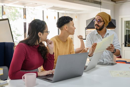 Three colleagues discussing work at a table with laptops in a bright office space.