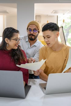 Diverse group of young adults working together, reviewing documents, and using laptops indoors.