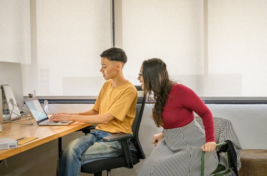 Two colleagues collaborating at a desk, engaging with a laptop in a modern office setting.