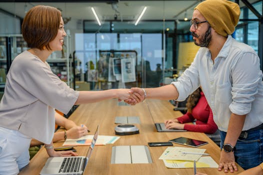 Two professionals shaking hands over a meeting table in a modern office setting.