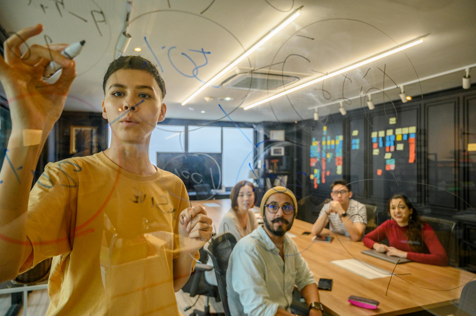 A team of young professionals working together in a modern office setting, brainstorming and writing on a glass board.