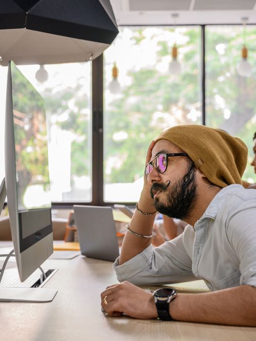 Free Bearded man with eyeglasses deep in thought while working at a computer in a modern office setting. Stock Photo