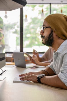 一名戴着无檐帽和眼镜的男子在明亮的办公室里对着电脑工作