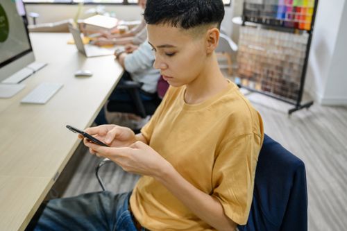 Free A young adult using a smartphone while working at a desk in a modern office setting. Stock Photo