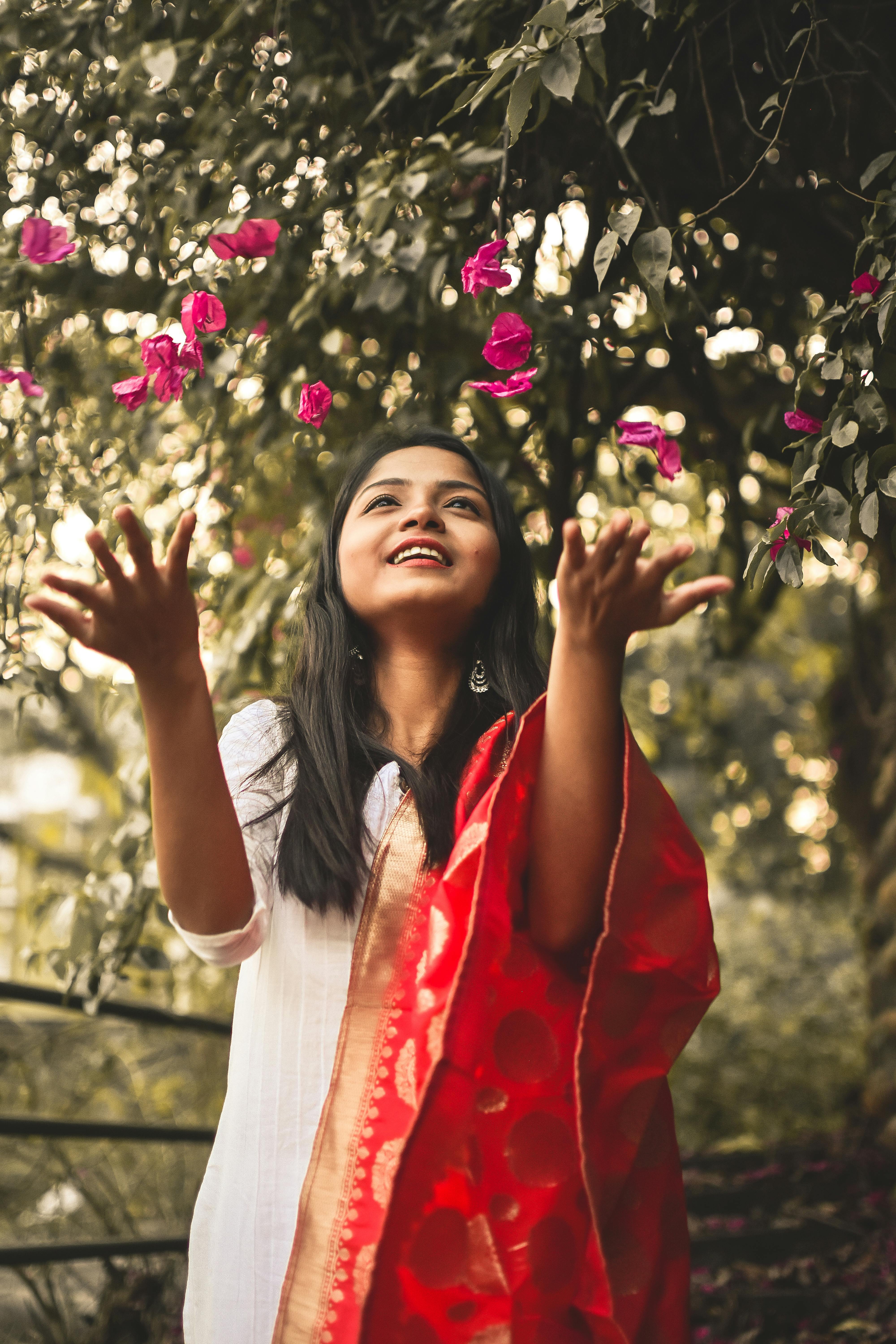 A smiling Indian woman in traditional attire reaching towards pink flowers, evoking a sense of joy.