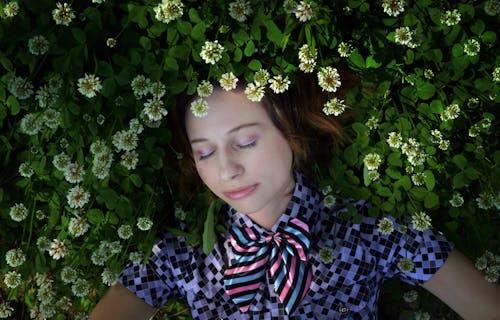 From above of young dreamy female in trendy clothes with soft makeup lying on green grass and small blossoming chamomiles