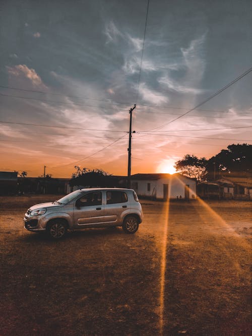 Silver automobile parked on dry grass field near suburban houses in village on sunset