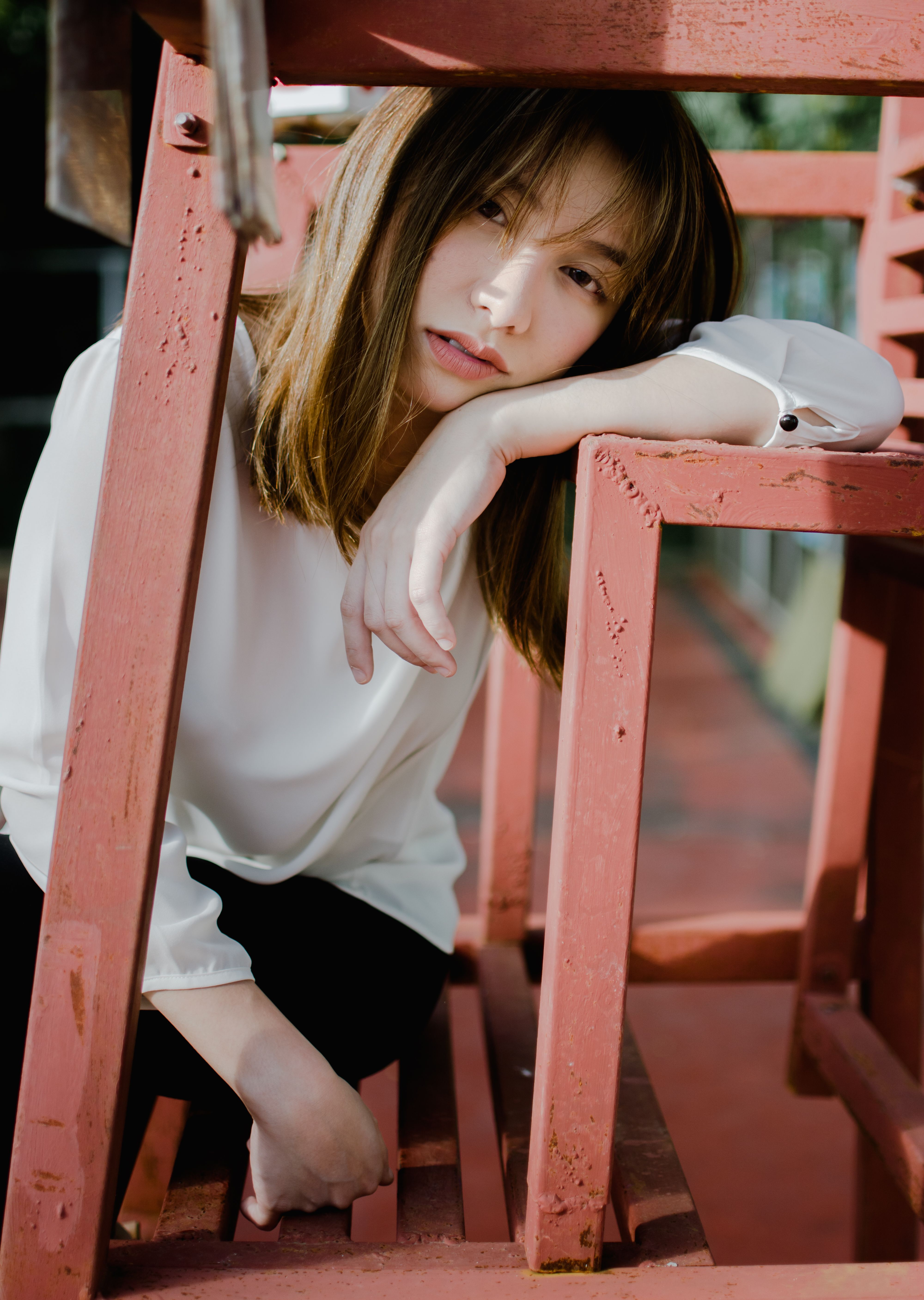 Bored young Asian female millennial in casual clothes sitting on metal bench and leaning on hand on sunny day