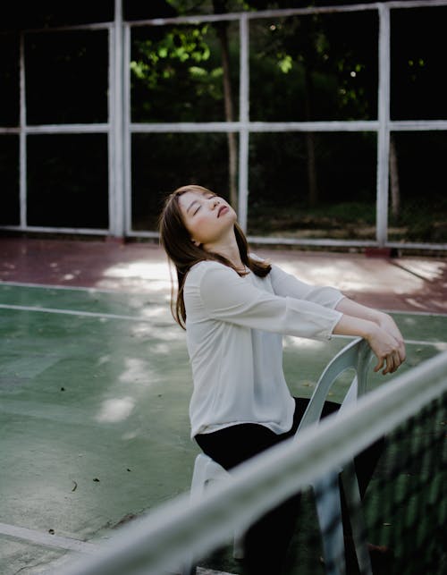 Dreamy young ethnic lady with dark hair in white shirt sitting on chair on tennis court with closed eyes