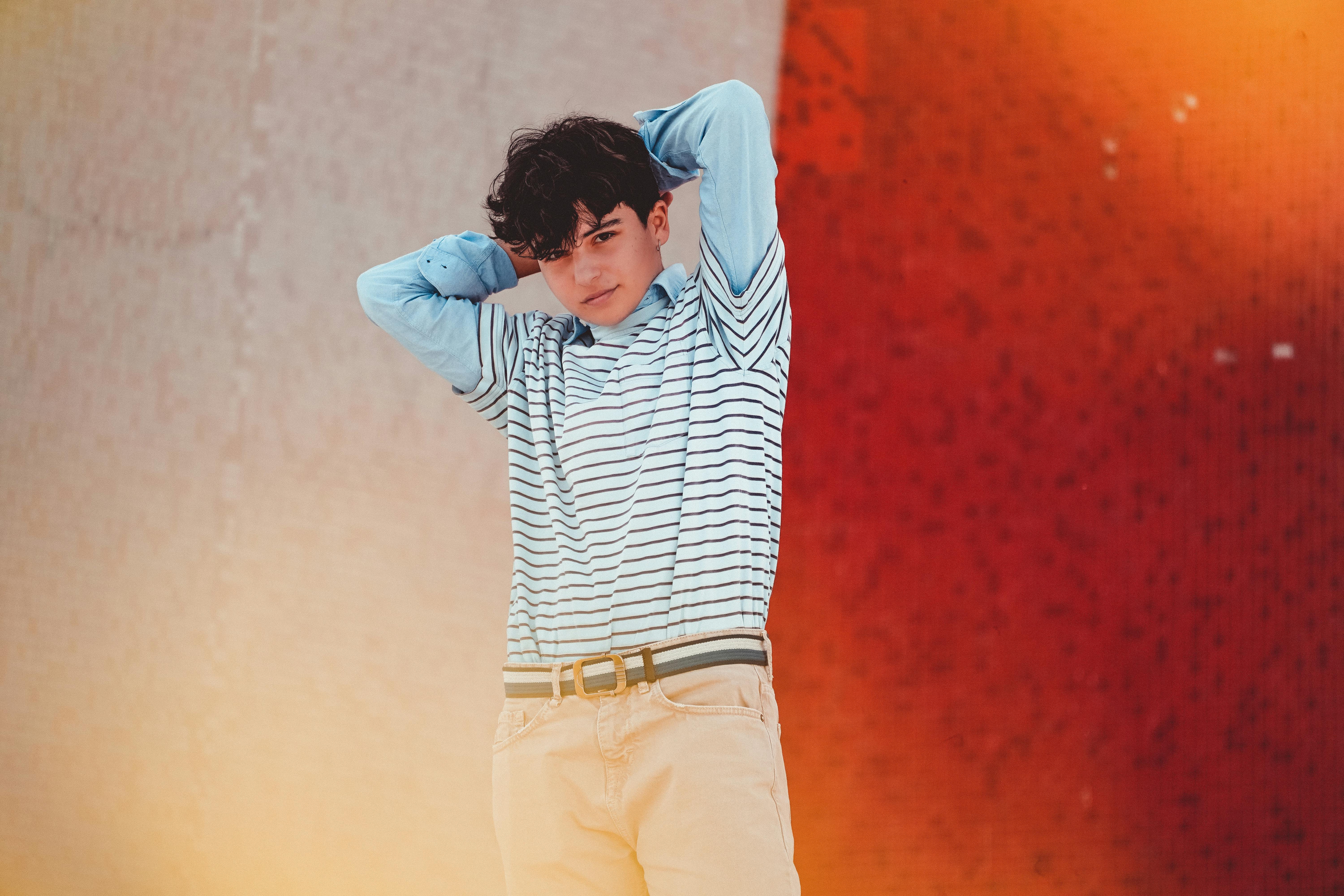 Content young guy in casual clothes standing on street near shabby wall with hands behind head and looking at camera