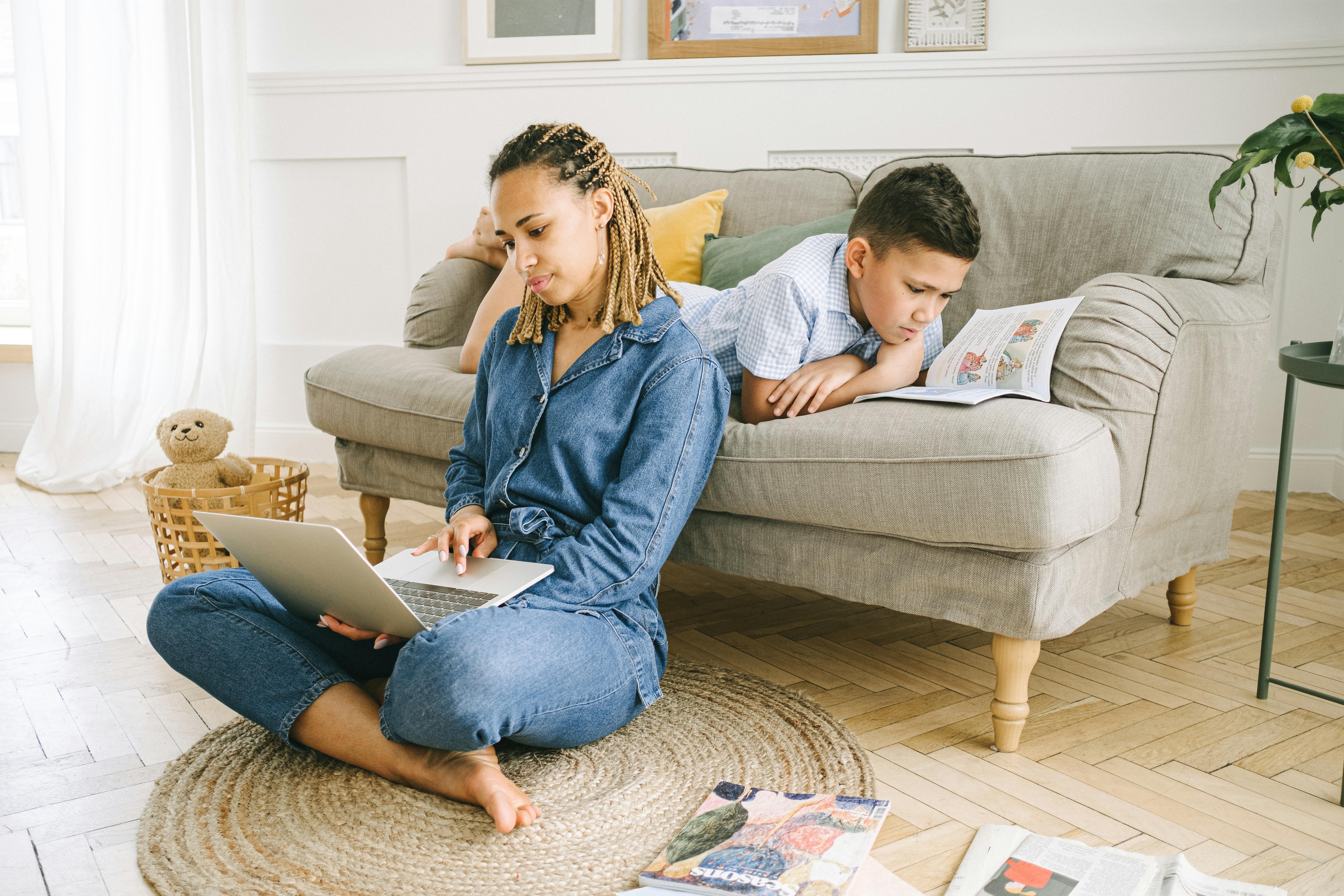 A mother working on a laptop while her son reads on the couch in a cozy living room.