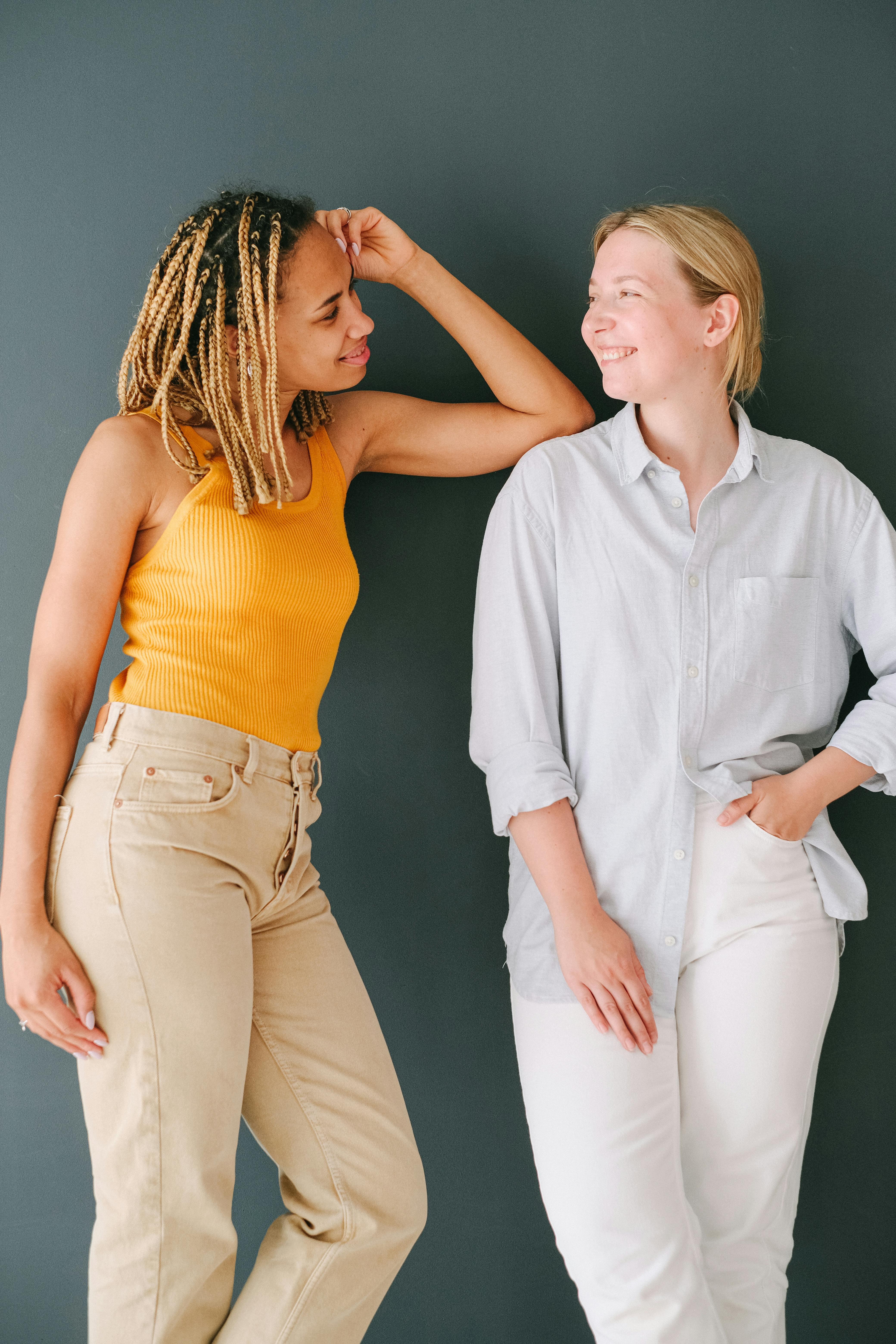 Two young women smiling and leaning casually against a wall indoors.