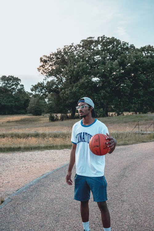 Free Contemplative African American male in eyeglasses and cap standing with basketball on roadway while looking away Stock Photo