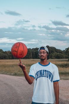 Young fit black male athlete in eyeglasses and sportswear spinning bright basketball on finger while looking at camera standing on road