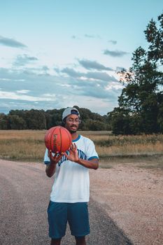 Young African American slim male in sports clothes and eyewear demonstrating basketball on roadway under sky and looking away
