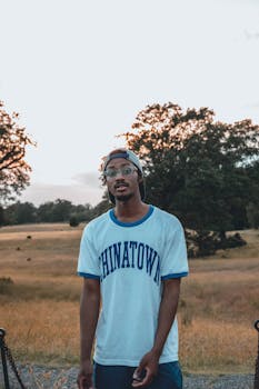 A young man stands thoughtfully in a rural field during dusk, surrounded by nature.